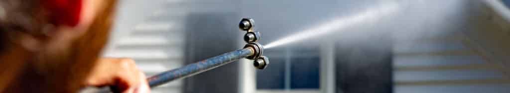 Power washing wand spraying white vinyl siding at a safe pressure to remove dirt and algae