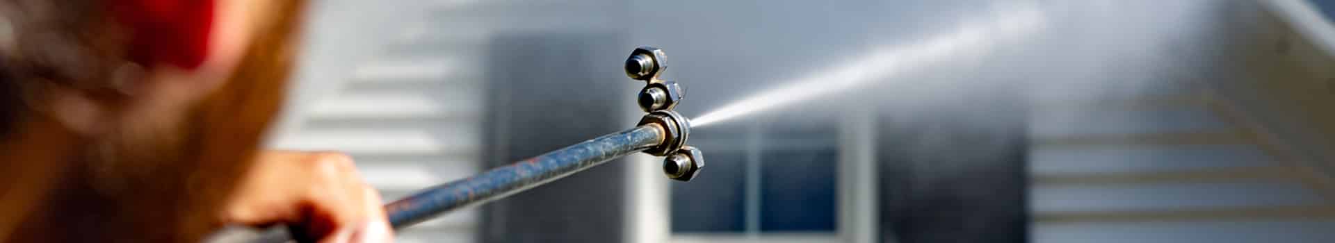 Power washing wand spraying white vinyl siding at a safe pressure to remove dirt and algae